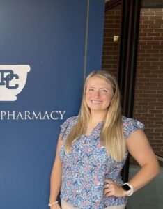 Current PCSP student, Audra Butler, beside a blue School of Pharmacy a wall 