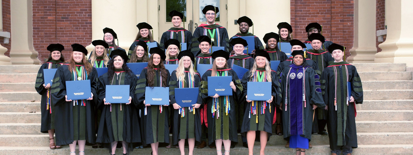 Class photo of the Class of 2025 Pharmacy Cohort in graduation regalia holding up their diplomas on the staircase in front of Neville